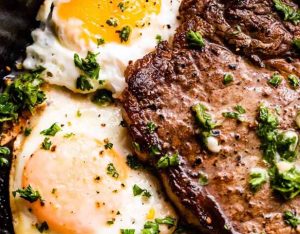 Sizzling sirloin steak and sunny-side-up eggs on a plate with fresh herbs.