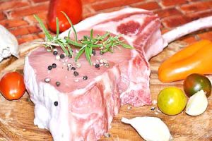Close-up of a raw, marbled Tomahawk Steak, seasoned with coarse salt and pepper, alongside fresh garlic cloves, ripe cherry tomatoes, and a chef's knife, ready for grilling on a Big Green Egg.