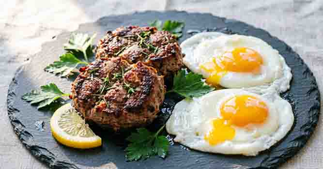Herbed lamb patties garnished with thyme and parsley served with fried eggs and a lemon wedge on dark slate.
