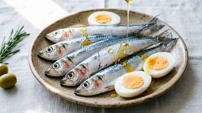 Whole silver sardines drizzled with golden olive oil alongside halved hard-boiled eggs on a speckled ceramic plate with rosemary.