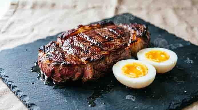 Juicy grilled ribeye steak resting on slate next to soft-boiled eggs cut in half, steam rising from the hot meat.