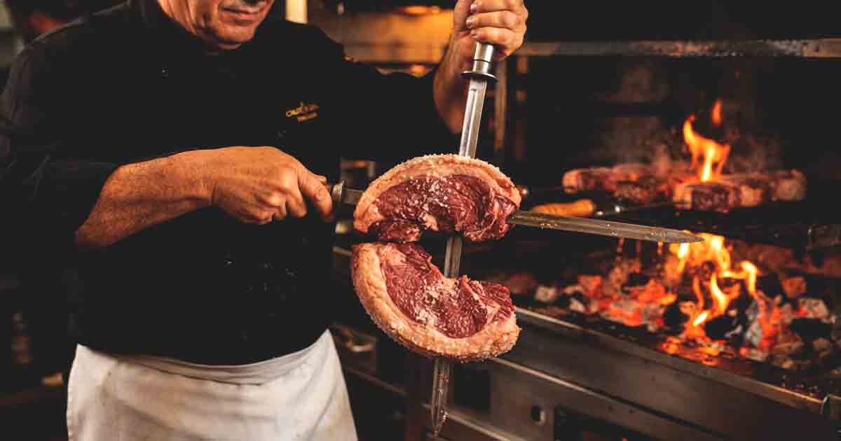 what is picanha, A bustling Brazilian steakhouse kitchen, where a gaucho chef, dressed in a black uniform and white apron, is carefully folding thick, raw picanha steaks into a 'C' shape and skewering them onto a long metal skewer.