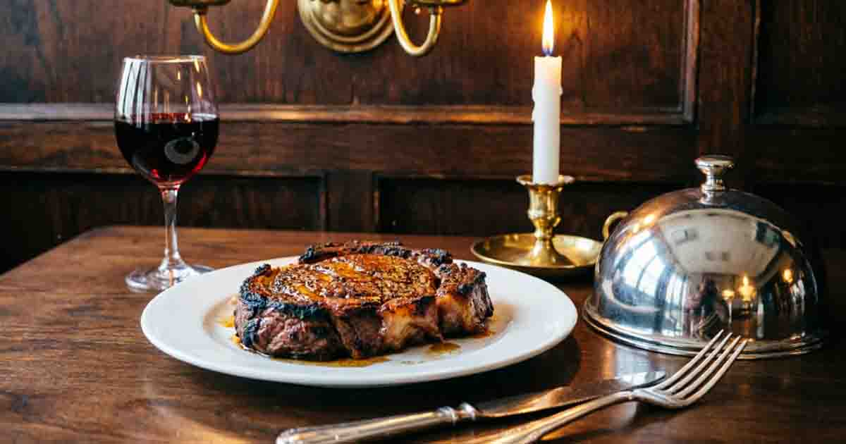 Classic steakhouse presentation of a pan-seared Delmonico steak on a white plate with red wine and candlelight.