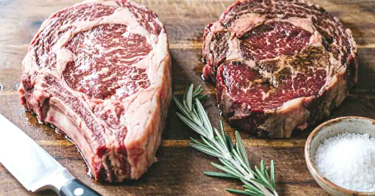Two raw, well-marbled Delmonico steak cuts on a wooden butcher block next to fresh rosemary and sea salt.