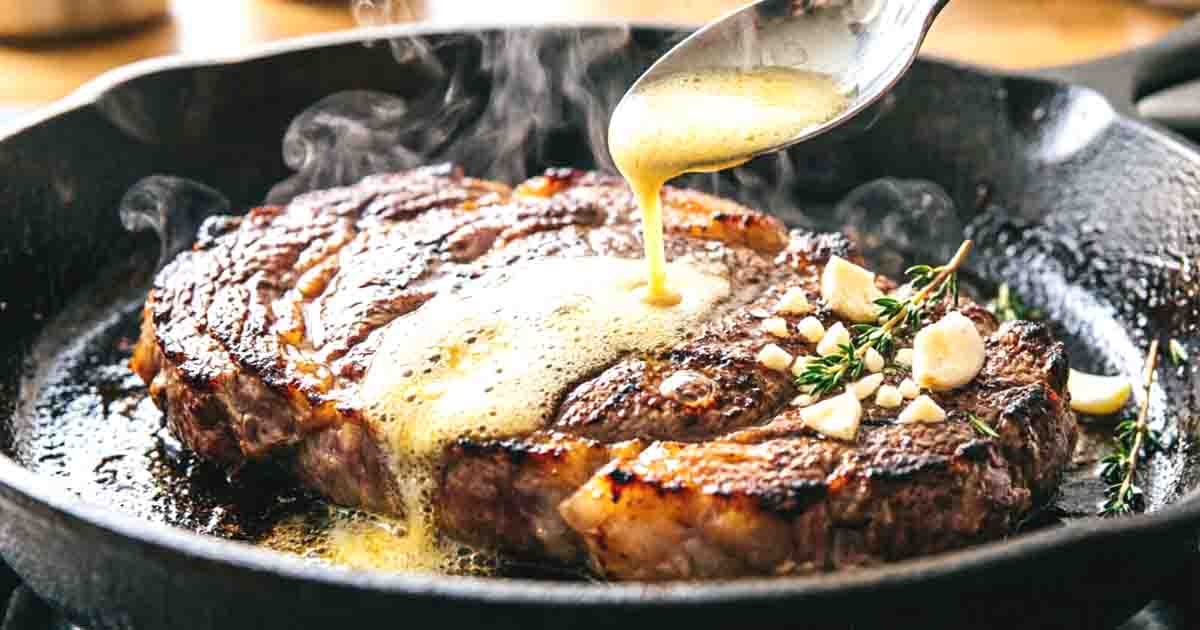 Basting a pan-seared Delmonico steak with melted garlic herb butter in a hot cast-iron skillet.