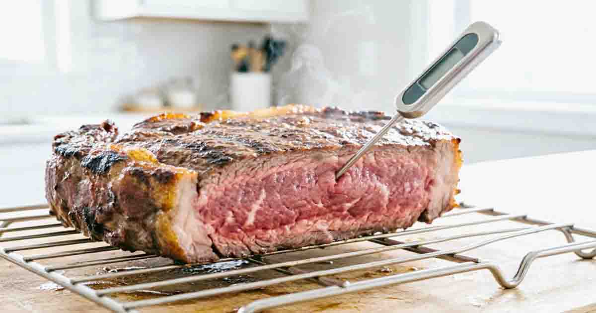 Digital meat thermometer measuring the internal temperature of a thick Delmonico steak on a wire cooling rack.