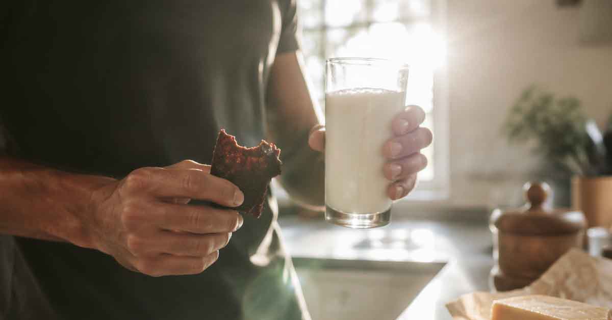 Healthy person holding a glass of cold whole milk and grass-fed beef jerky in a sunlit modern farmhouse kitchen, representing clean carnivore diet snack choices.