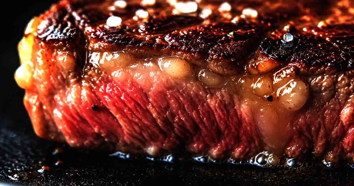 carnivore diet, Close-up macro view of a sizzling medium-rare ribeye steak showing marbled rendered fat and crispy seared crust.