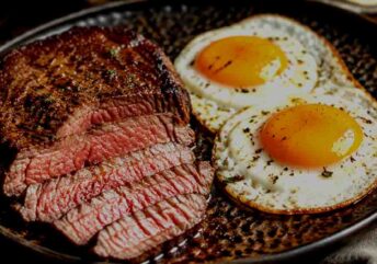 Pan-seared medium-rare ribeye steak served with two sunny-side-up eggs in a cast-iron skillet for a high-protein keto breakfast. Love your ribeye steak and eggs meal